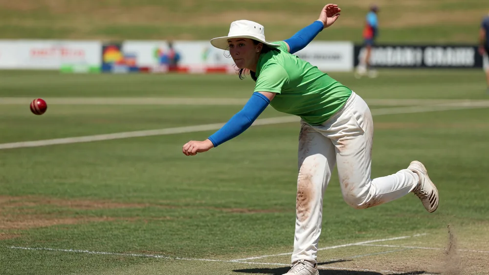 Woman pace bowler in a green shirt, white hat and blue sleeves releasing a red cricket ball on a sunny ground
